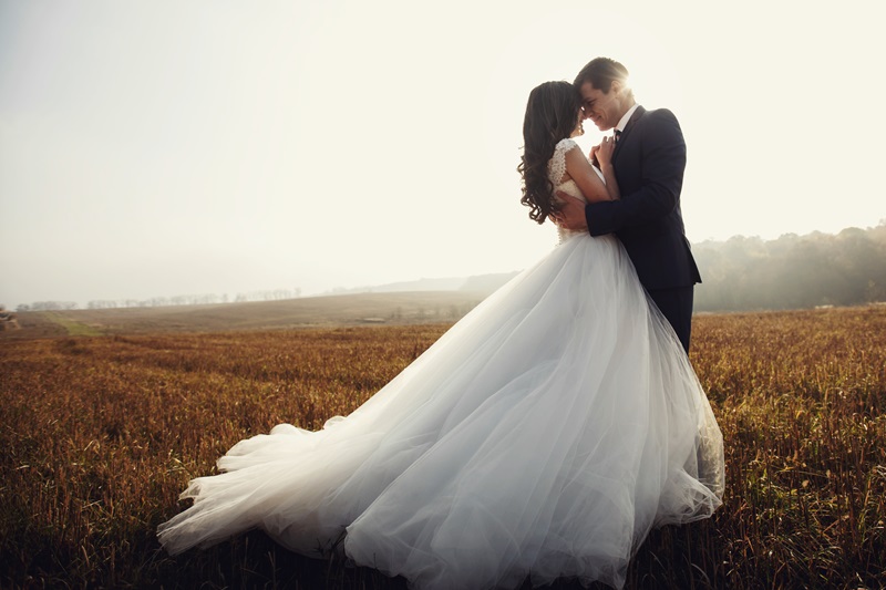 Romantic fairytale newlywed couple hug &amp; kiss in field at sunset